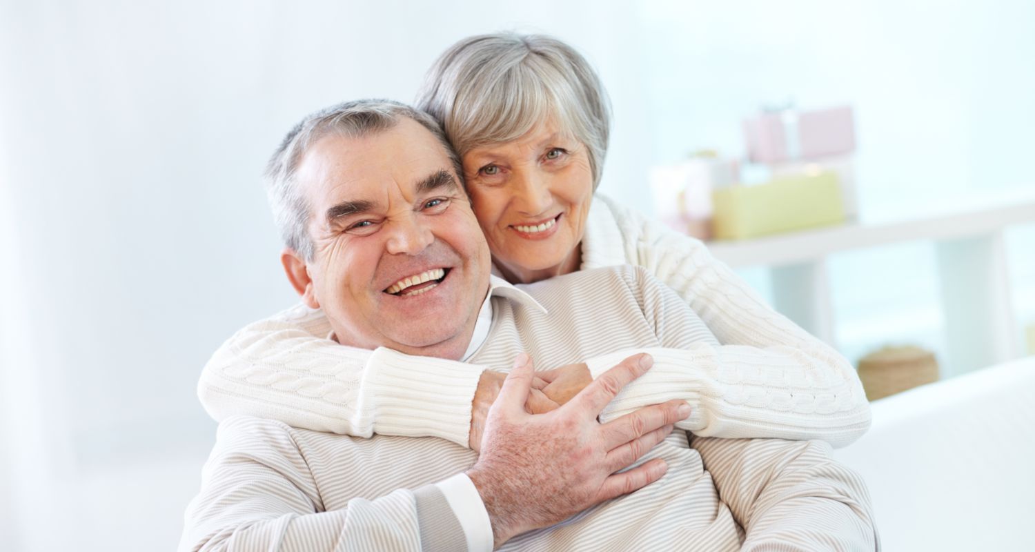 Elderly couple smiling with wife's arms around husband